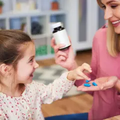 Uśmiechnięta mama podaje dziecku niebieskie kapsułki. Sprawdź, jak podać dziecku antybiotyk, żeby nie wypluło.