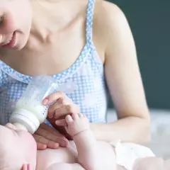 Uśmiechnięta mama karmi butelką noworodka. Warto wiedzieć, ile powinien jeść dwutygodniowy noworodek, by zapewnić mu prawidłowy rozwój.