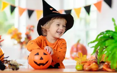 Uśmiechnięty chłopiec w czarnej, spiczastej czapce czarownicy i pomarańczowej bluzce z nietoperzami trzyma dynię. To idealny strój owoc dla dziecka na Halloween!