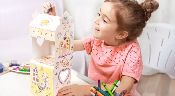 Uśmiechnięta dziewczynka ozdabia kartonowy domek, który jest świetną zabawka dla sześciolatków. Kolorowe kredki i farby czekają na użycie.