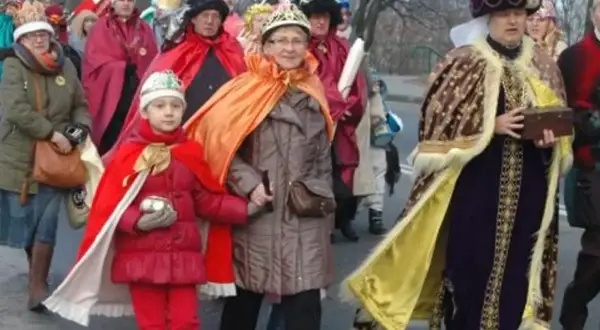 Kolorowe stroje Trzech Króli na ulicznym orszaku. Dziecko w czerwonym płaszczu i koronie, obok dorosłe osoby w bogatych szatach.