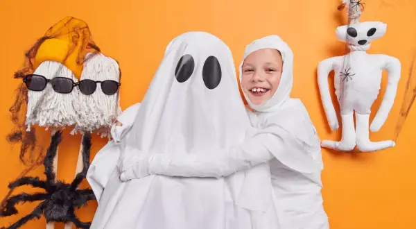 Dzieci w strojach mumii i ducha, świetny pomysł, jak zrobić strój na Halloween. Obok pluszowy szkielet i pająk.