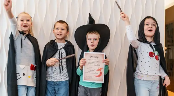 Cztery dzieci w strojach czarodziejów z różdżkami. Jeden chłopiec w kapeluszu czarownicy trzyma dyplom.