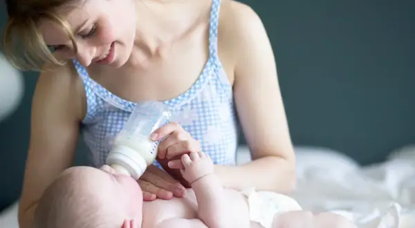 Uśmiechnięta mama karmi butelką noworodka. Warto wiedzieć, ile powinien jeść dwutygodniowy noworodek, by zapewnić mu prawidłowy rozwój.